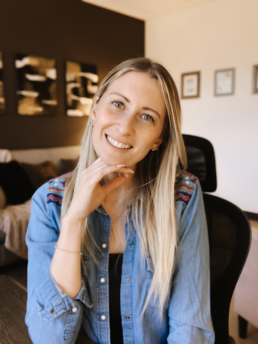 Smiling blonde woman with blue eyes, wearing a denim shirt, resting her chin on her hand in an office setting.