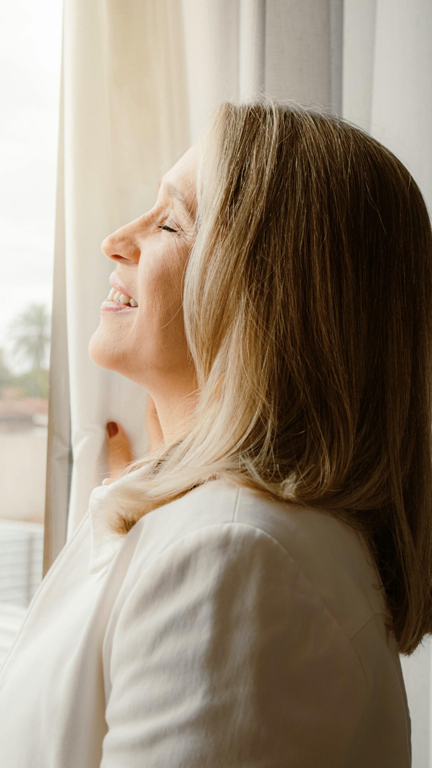 Blonde, mature woman, standing at the window, eyes closed, sun on her face, smiling, therapyworthwhile