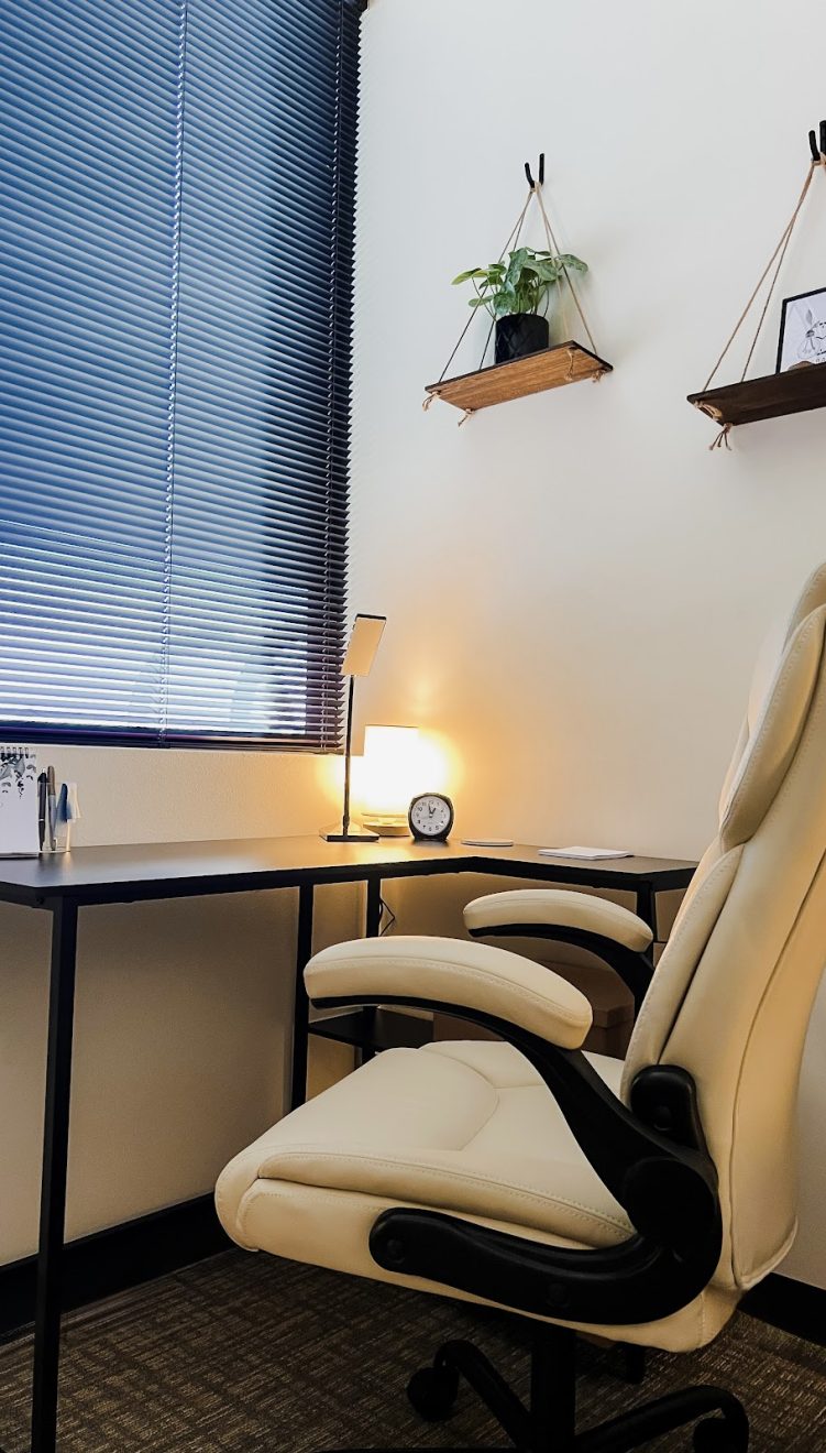 A cream-colored ergonomic office chair facing a minimalist L-shaped desk with a desk calendar, lamp, and hanging wooden shelves.