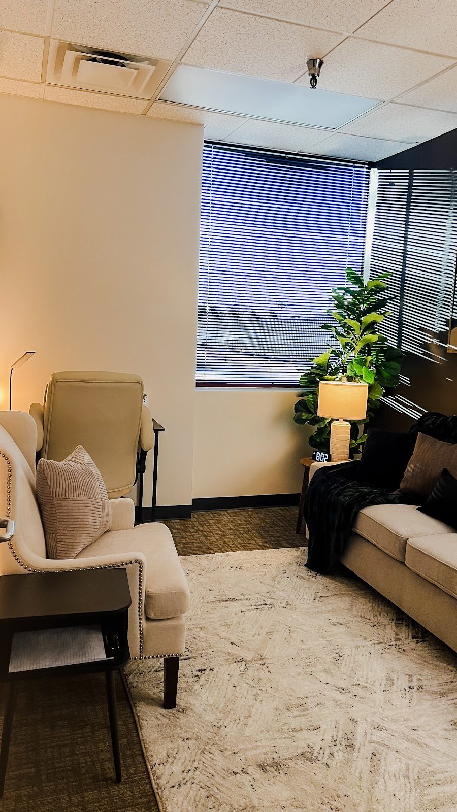 Interior view of a cozy therapy office featuring a cream sofa, a matching armchair, a patterned rug, and soft lamp lighting.